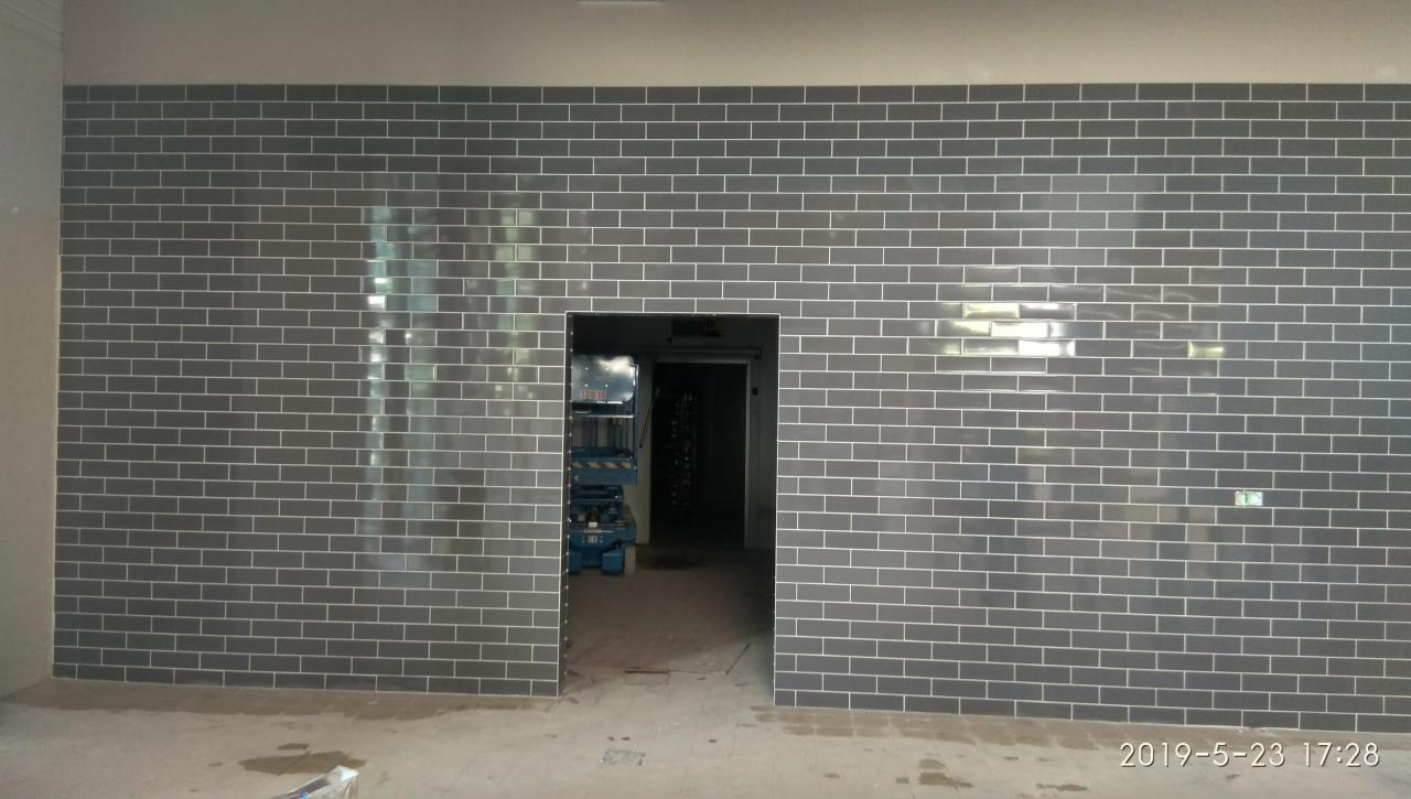 Ściana wyłożona błyszczącymi, szarymi płytkami imitującymi cegłę, z widocznym otworem drzwiowym i podnośnikiem nożycowym w tle, wykonana w Obornikach.