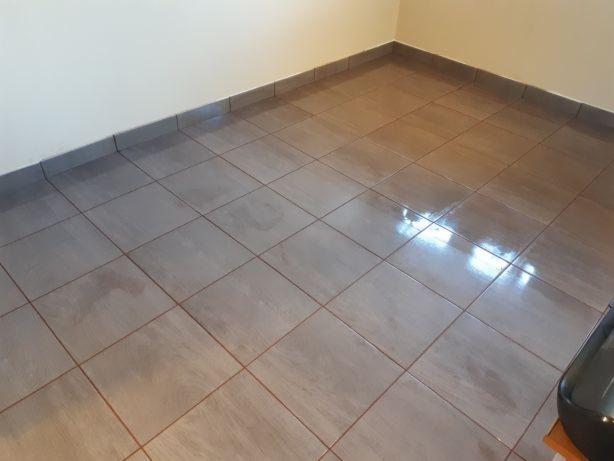 Wykończona podłoga z szarych, kwadratowych płytek ceramicznych, z ciemnoszarą listwą przypodłogową przy jasnej ścianie.