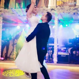 Para młoda tańczy podczas wesela, on podnosi ją w górę, w tle zespół muzyczny i goście na balkonie.