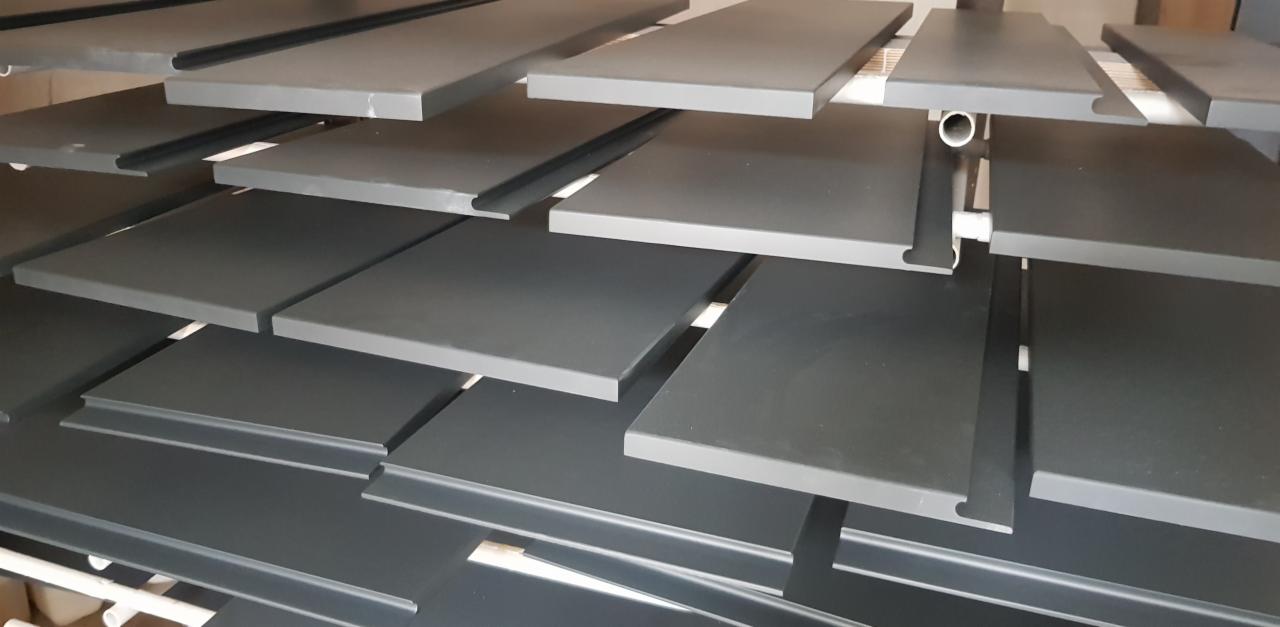 Szare, pomalowane elementy drewniane, prawdopodobnie blaty lub półki, suszone na stojaku w lakierni.