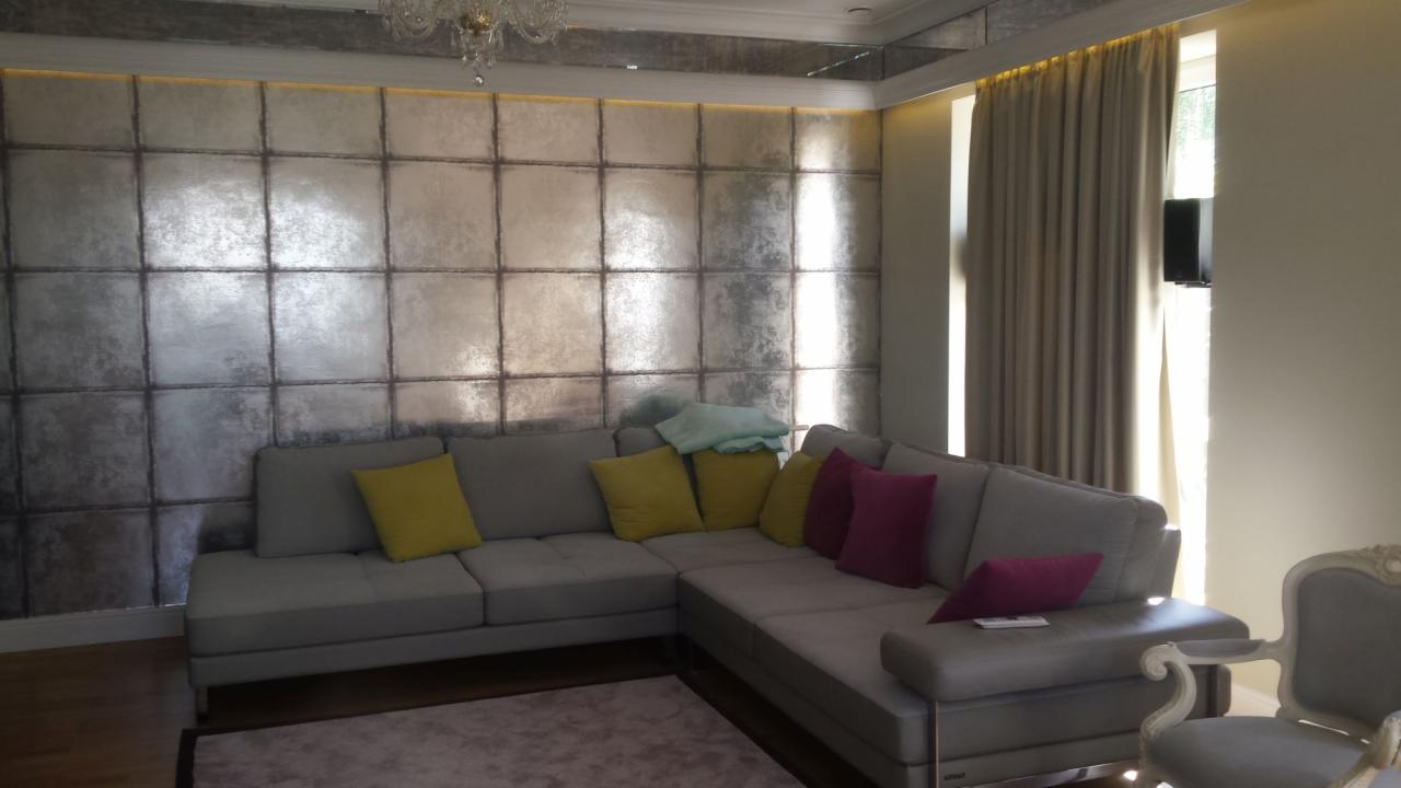 Salon z narożną sofą na tle ściany wykończonej panelami imitującymi metalowe płyty, oświetlonej od góry, z lustrzanym sufitem i żyrandolem.