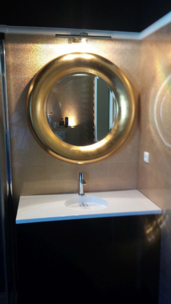 Okrągłe lustro w złotej ramie nad białą umywalką z chromowaną baterią, zamontowaną na czarnej szafce w łazience z beżowymi płytkami i oświetleniem punktowym.