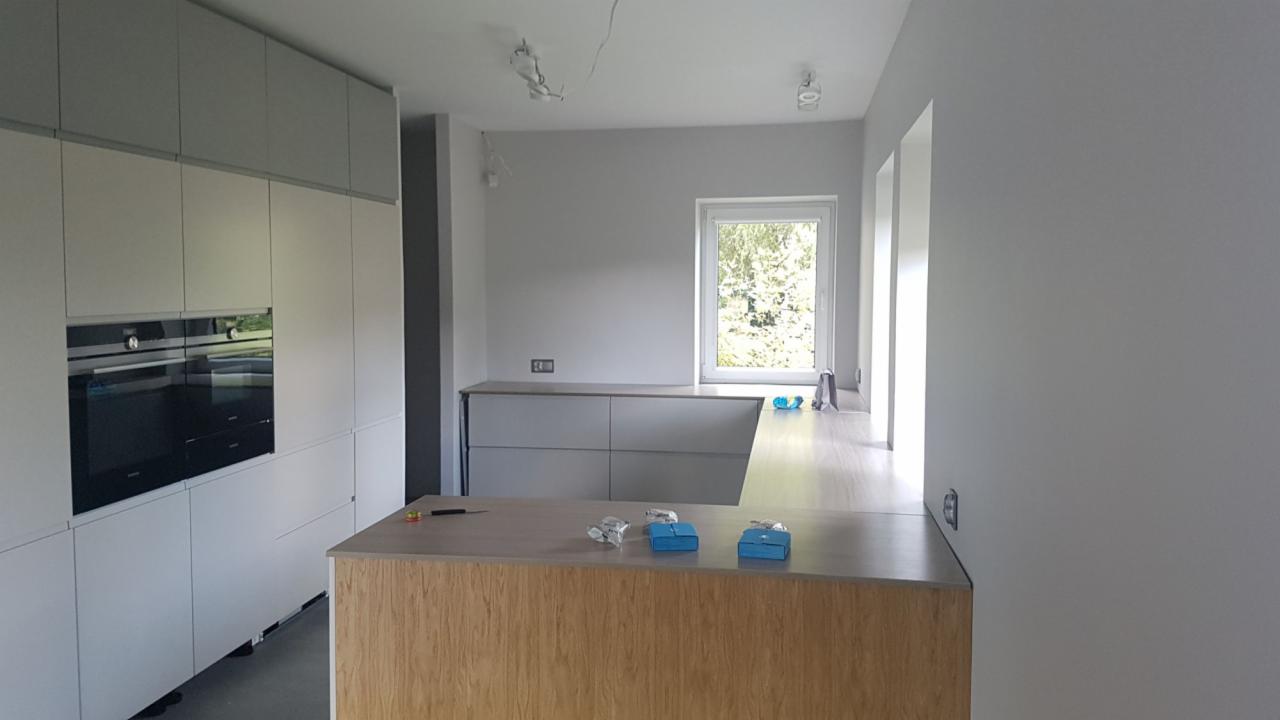 Jasna kuchnia w trakcie montażu: szare szafki górne i dolne, drewniany półwysep z narzędziami i niebieskimi pudełkami, okno z widokiem na zieleń.