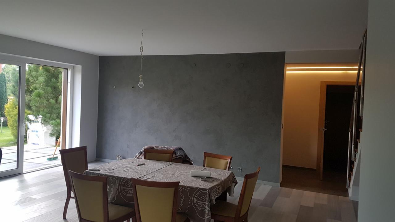 Pokój z szarą ścianą o fakturze betonu, nakryty stół i niedokończone oświetlenie sufitowe, widoczne ślady po demontażu lampy.