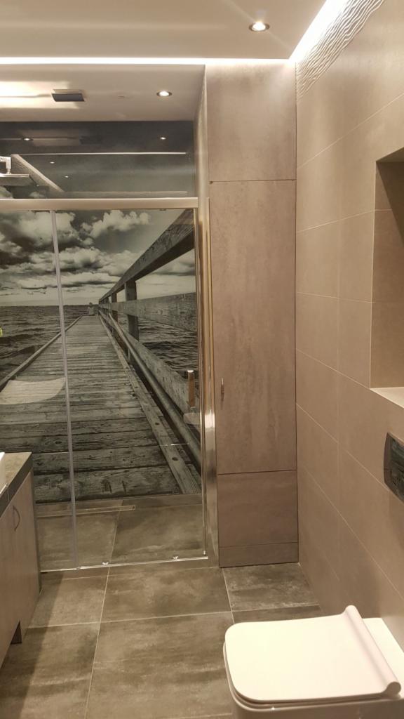Nowoczesna łazienka z kabiną prysznicową z fototapetą przedstawiającą drewniany pomost nad morzem, beżowe płytki na ścianach i podłodze, oraz zabudowana szafka. Minimalistyczna toaleta w prawym...