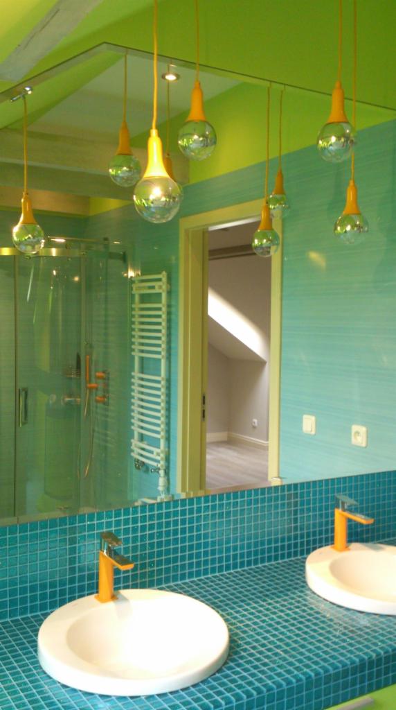 Turkusowa mozaika na blacie umywalkowym z dwoma białymi umywalkami i pomarańczowymi bateriami, widok na kabinę prysznicową i grzejnik drabinkowy, wiszące lampy w kształcie żarówek i zielone ściany.