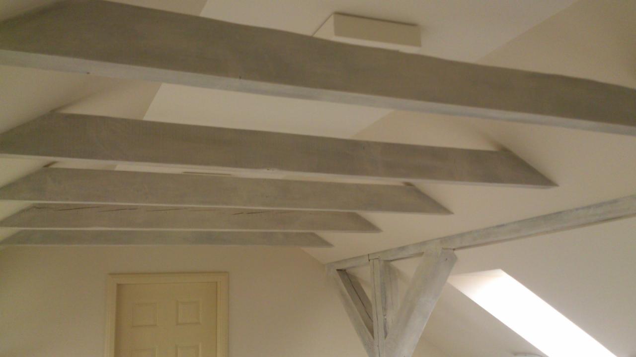 Wykończone poddasze z drewnianymi belkami stropowymi pomalowanymi na szaro, widoczne białe ściany i fragment okna dachowego.