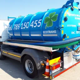 Ciężarówka asenizacyjna z niebieskim zbiornikiem ozdobionym logo firmy Ecotrans w kształcie zielonej koniczyny i numerem telefonu, zaparkowana na betonowym placu w słoneczny dzień.