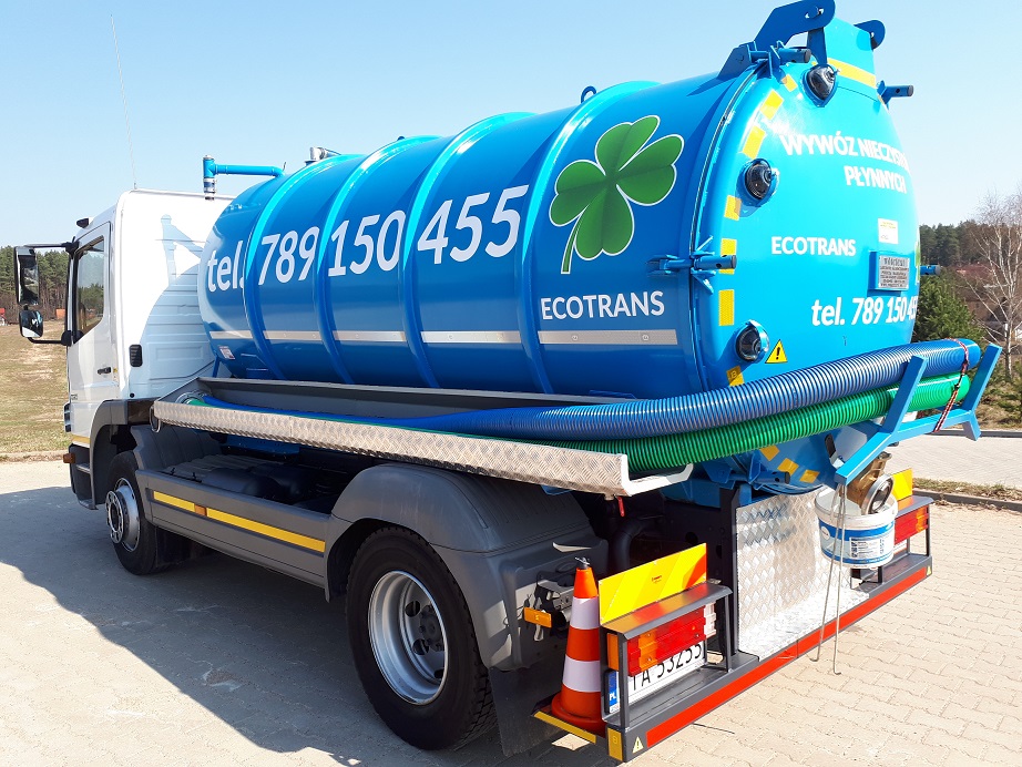 Ciężarówka asenizacyjna z niebieskim zbiornikiem ozdobionym logo firmy Ecotrans w kształcie zielonej koniczyny i numerem telefonu, zaparkowana na betonowym placu w słoneczny dzień.
