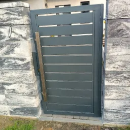 Nowoczesna, pozioma brama wjazdowa w kolorze grafitowym, zamontowana między dwoma słupkami z szarej kostki imitującej kamień.