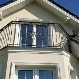 Elegancki balkon z czarną, metalową balustradą o ozdobnym wzorze, umieszczony na piętrze domu o jasnej elewacji i ciemnej dachówce.