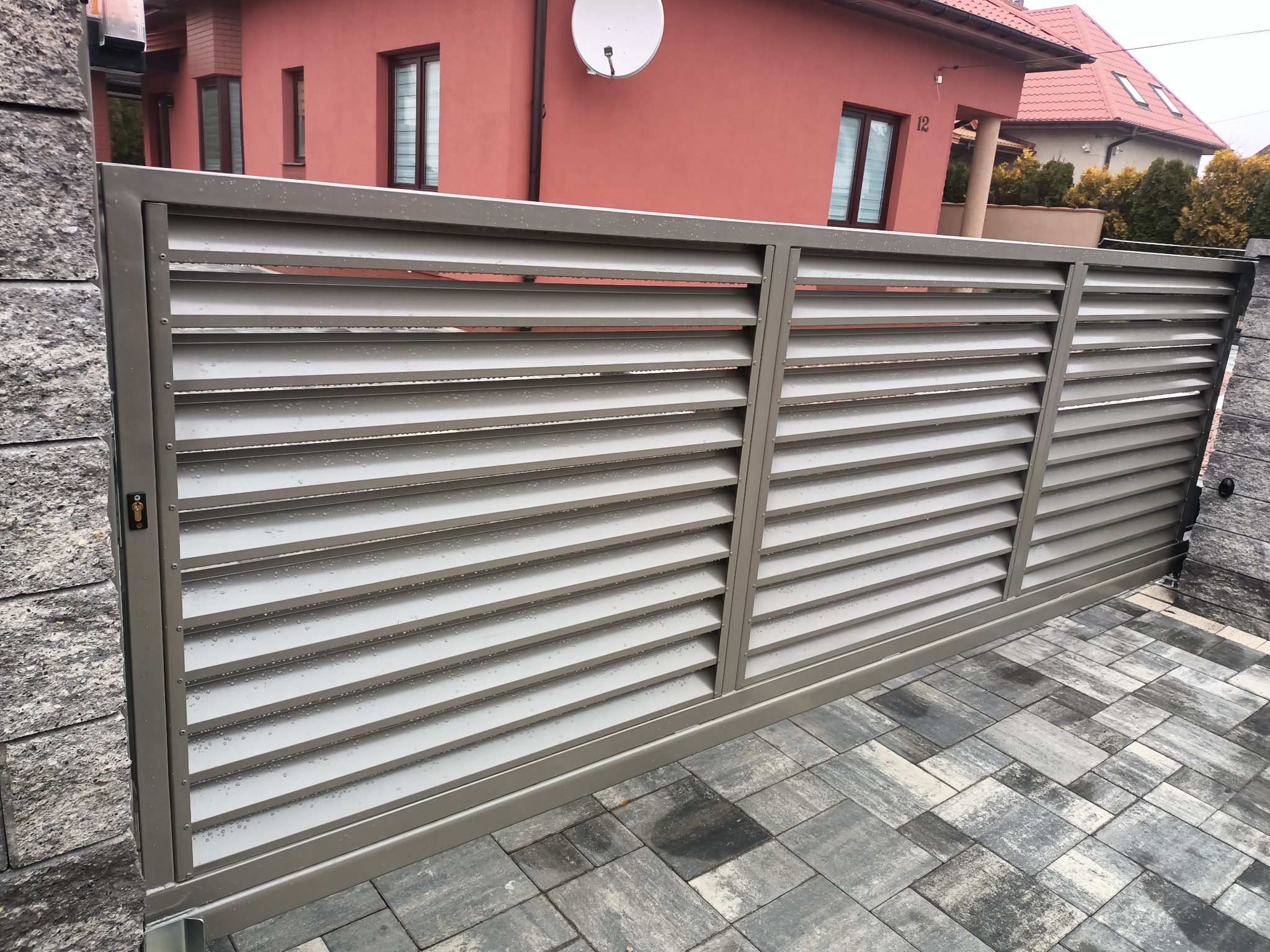 Przesuwna, pozioma brama lamelowa w kolorze szarym, z widocznymi kroplami deszczu, zamontowana przed domem z elewacją w odcieniach czerwieni.