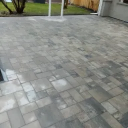 Ułożona kostka brukowa w odcieniach szarości i brązu tworząca wzór na tarasie przy domu z widokiem na trawnik i drzewa.