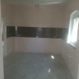 Przygotowane do wykończenia wnętrze kuchni z ułożonymi ciemnymi płytkami nad blatem, surowa podłoga, okno w łukowej wnęce, widoczne instalacje elektryczne i rury.