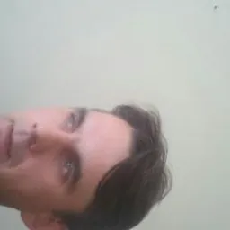 Portret mężczyzny z krótkimi, ciemnymi włosami i niebieskimi oczami, na jasnym tle. Ujęcie z dołu, lekko obrócone.