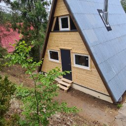 WOLKAM Kamil Wolski - Nowoczesny, drewniany domek letniskowy w kształcie litery A z szarą blachodachówką, czarnymi drzwiami i kominem ze stali nierdzewnej, otoczony drzewami i krzewami.