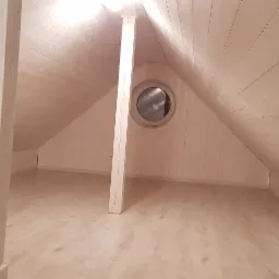 Wnętrze poddasza wykończonego w drewnie, z widoczną drewnianą podłogą, ścianami i sufitem, podtrzymywanym przez dwie drewniane kolumny oraz okrągłym oknem.