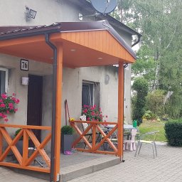 WOLKAM Kamil Wolski - Zadaszone wejście do domu z drewnianą balustradą w kolorze pomarańczowym, ozdobione kwiatami w oknach, widok na ogród z tyłu.