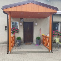 WOLKAM Kamil Wolski - Zadaszone wejście do domu z drewnianą konstrukcją, numerem 76 nad drzwiami, kwiatami w oknach i roślinami w donicach po bokach.