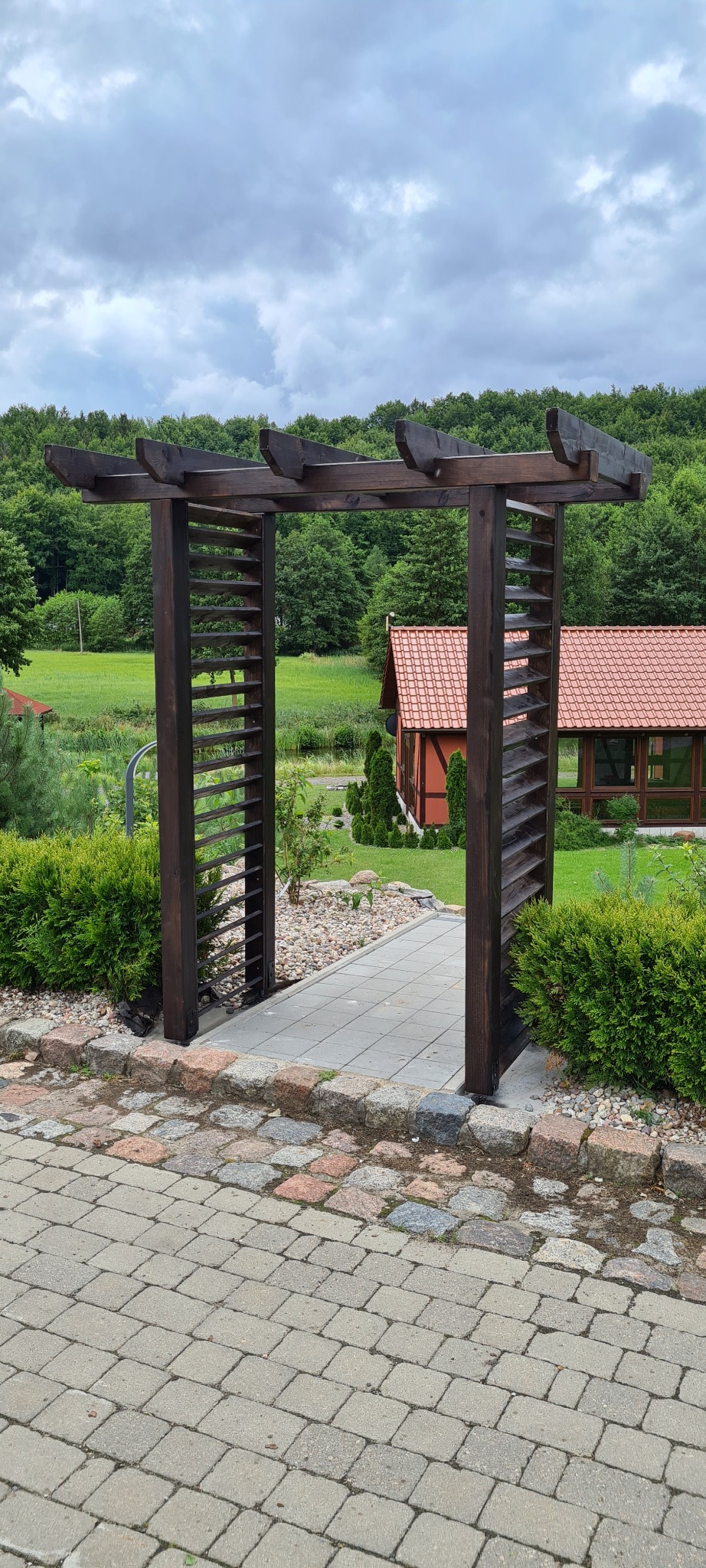 Ciemna, drewniana konstrukcja z listew tworząca wejście do ogrodu, z widokiem na dom z czerwoną dachówką w tle.