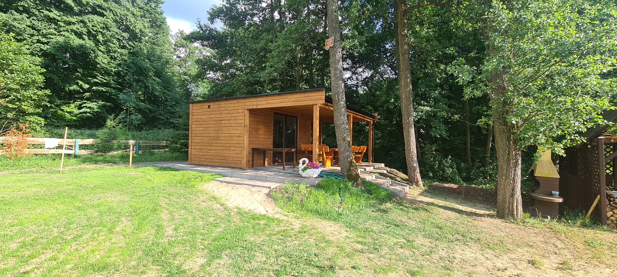 Nowoczesna, drewniana konstrukcja z płaskim dachem, pełniąca funkcję letniskowej altany, z drewnianymi meblami i dekoracyjną donicą w kształcie łabędzia, otoczona zielenią i drzewami.