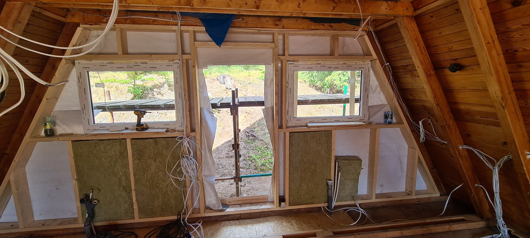Wnętrze poddasza w trakcie adaptacji: widoczne odeskowanie, drewniana konstrukcja, okna PCV, częściowo ułożona wełna mineralna, instalacja elektryczna w peszlach, narzędzia i materiały budowlane.