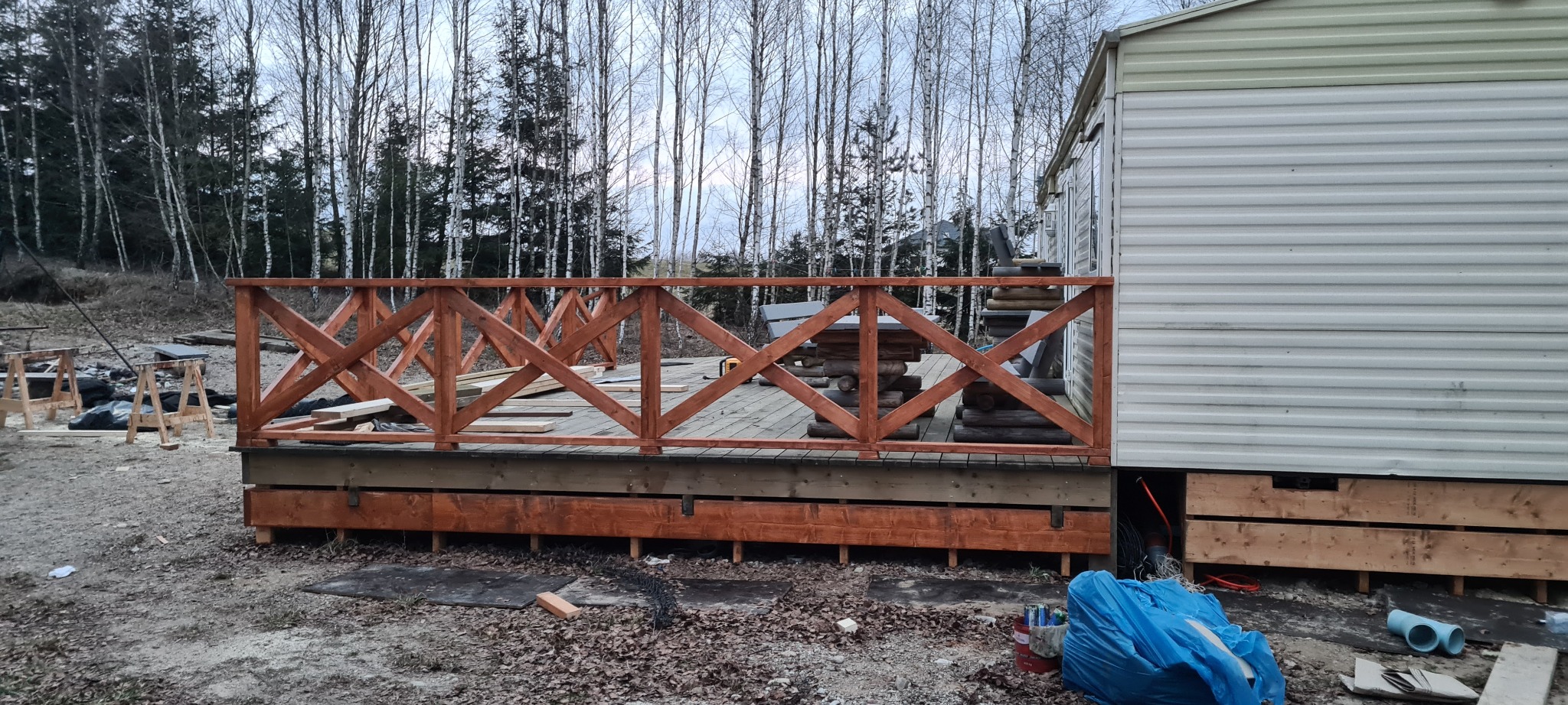 Drewniana balustrada z krzyżującymi się belkami pomalowana na brązowo, otaczająca taras przy domku mobilnym, widoczne narzędzia i materiały budowlane.