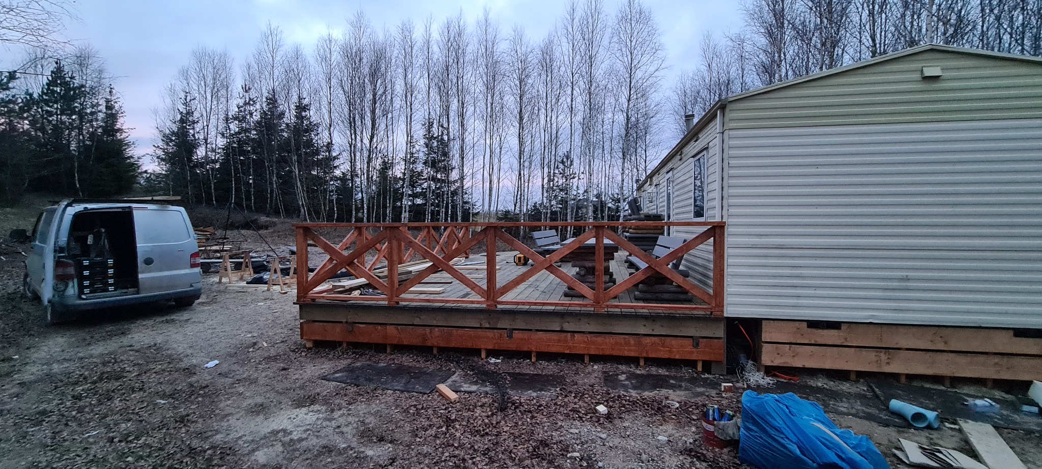 Budowa drewnianej balustrady z krzyżowym wzorem na tarasie przy domku letniskowym, widoczny samochód dostawczy z otwartymi drzwiami i narzędziami w tle lasu.