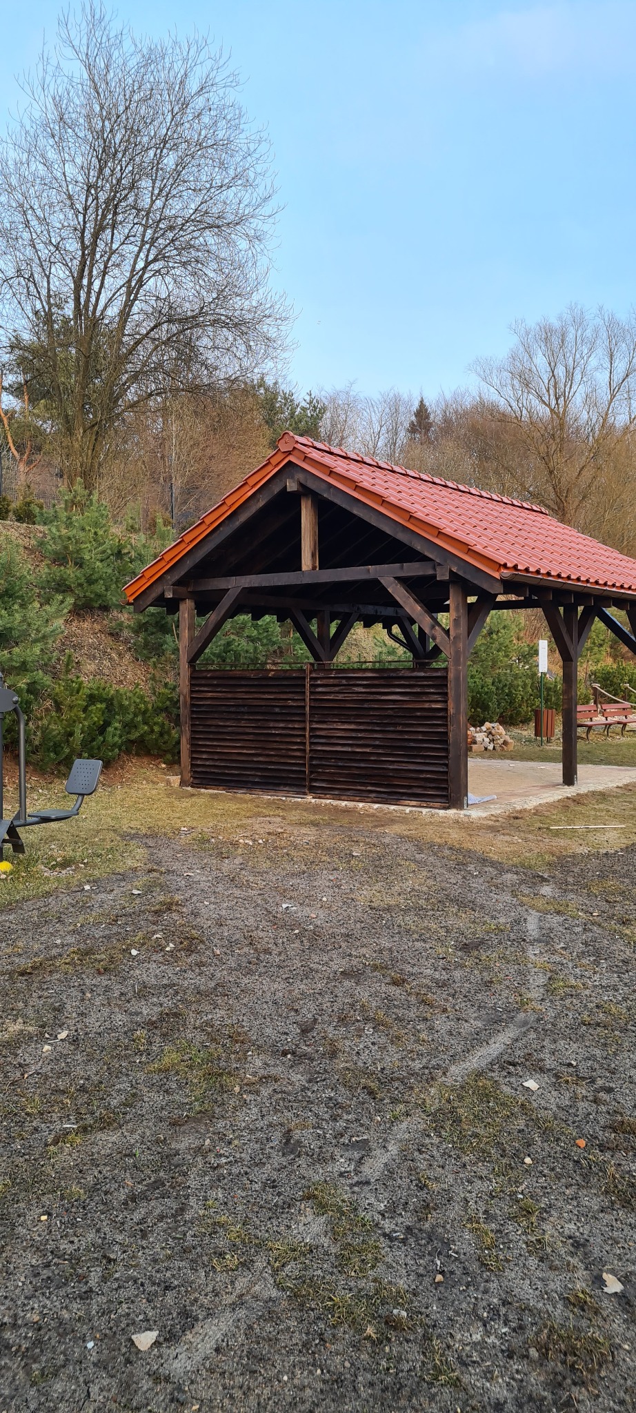 Drewniana altana z czerwonym dachem w parku, częściowo osłonięta poziomymi deskami, z widocznym sprzętem do ćwiczeń na pierwszym planie i drzewami w tle.