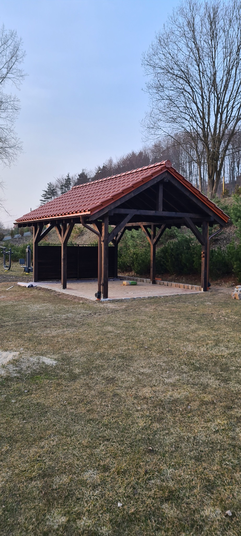 Zadaszona konstrukcja ogrodowa z ciemnego drewna i czerwoną dachówką, widoczna z perspektywy trawnika, z elementami placu zabaw w tle.