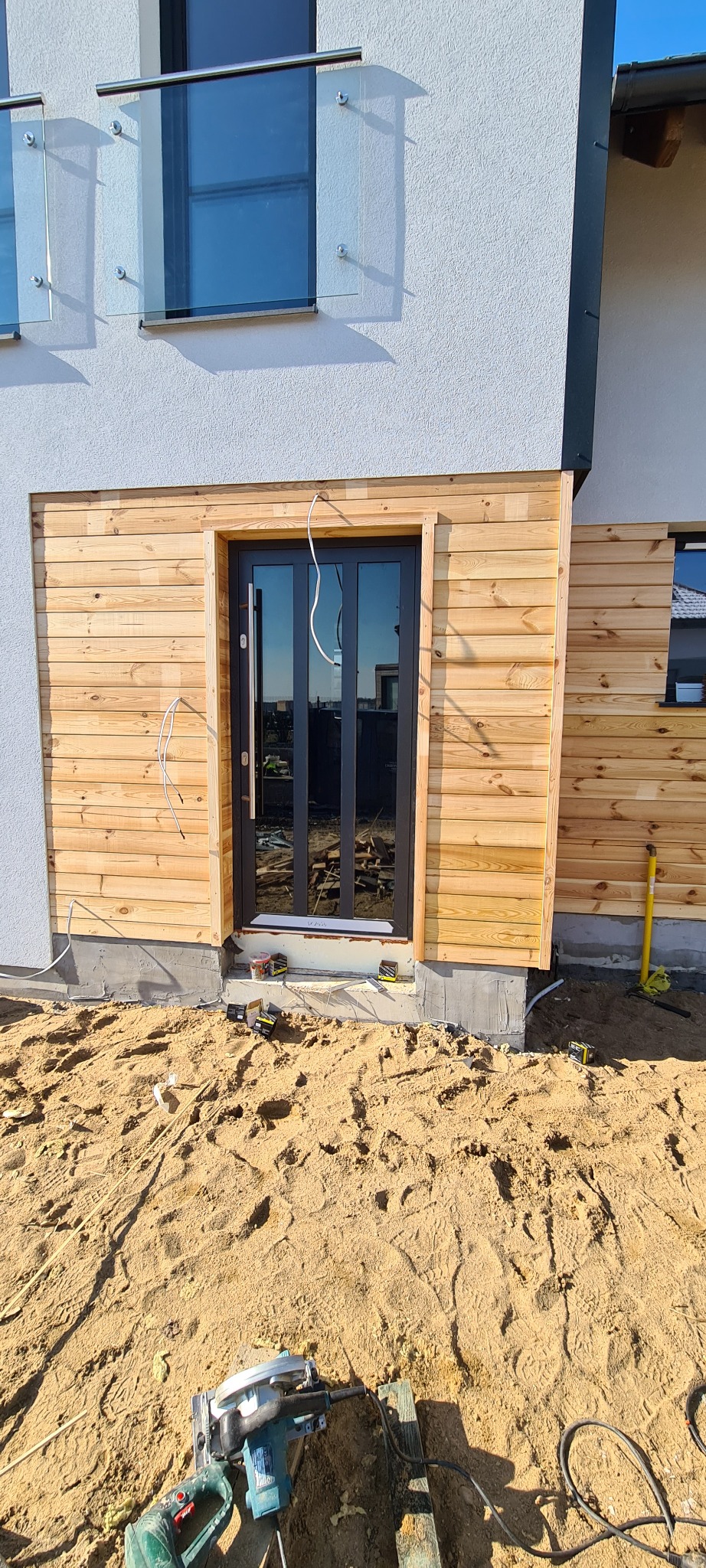 Montaż drewnianej elewacji wokół czarnych, szklanych drzwi tarasowych w nowym budynku; widoczne narzędzia i kable elektryczne na piaszczystym podłożu.