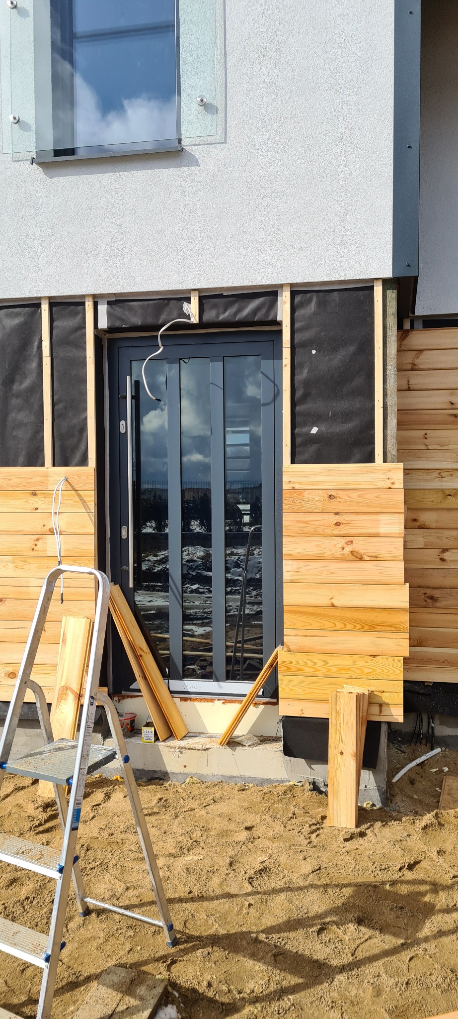 Montaż drewnianej elewacji na ścianie domu, widoczne deski, drabina i czarna membrana izolacyjna, w tle szare drzwi z szybami.