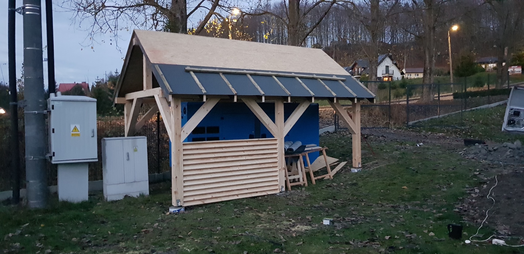 Wznoszona konstrukcja z drewna, prawdopodobnie altana lub wiata; widoczne elementy dachu z desek i papy, częściowo obudowane ściany z poziomych desek, w tle domy i drzewa.