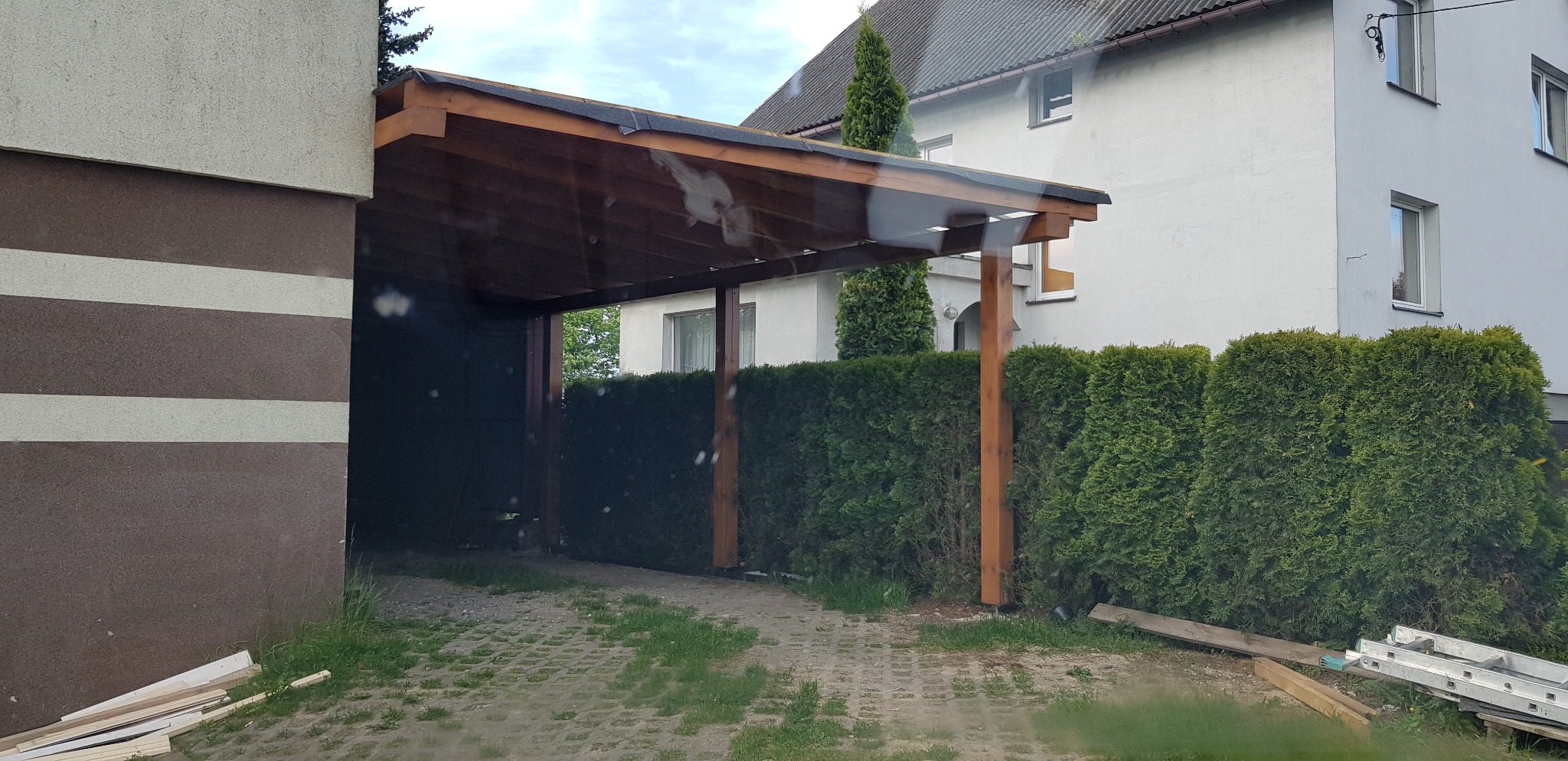 Drewniana wiata garażowa z płaskim dachem pokrytym czarną papą, wsparta na solidnych, brązowych słupach, przylegająca do budynku z brązowo-białą elewacją. W tle widoczny budynek mieszkalny...