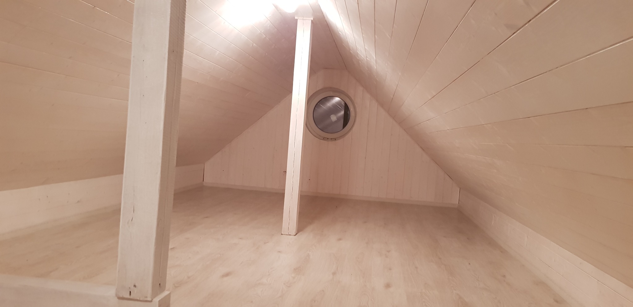 Wnętrze poddasza wykończonego w drewnie, z widoczną drewnianą podłogą, ścianami i sufitem, podtrzymywanym przez dwie drewniane kolumny oraz okrągłym oknem.
