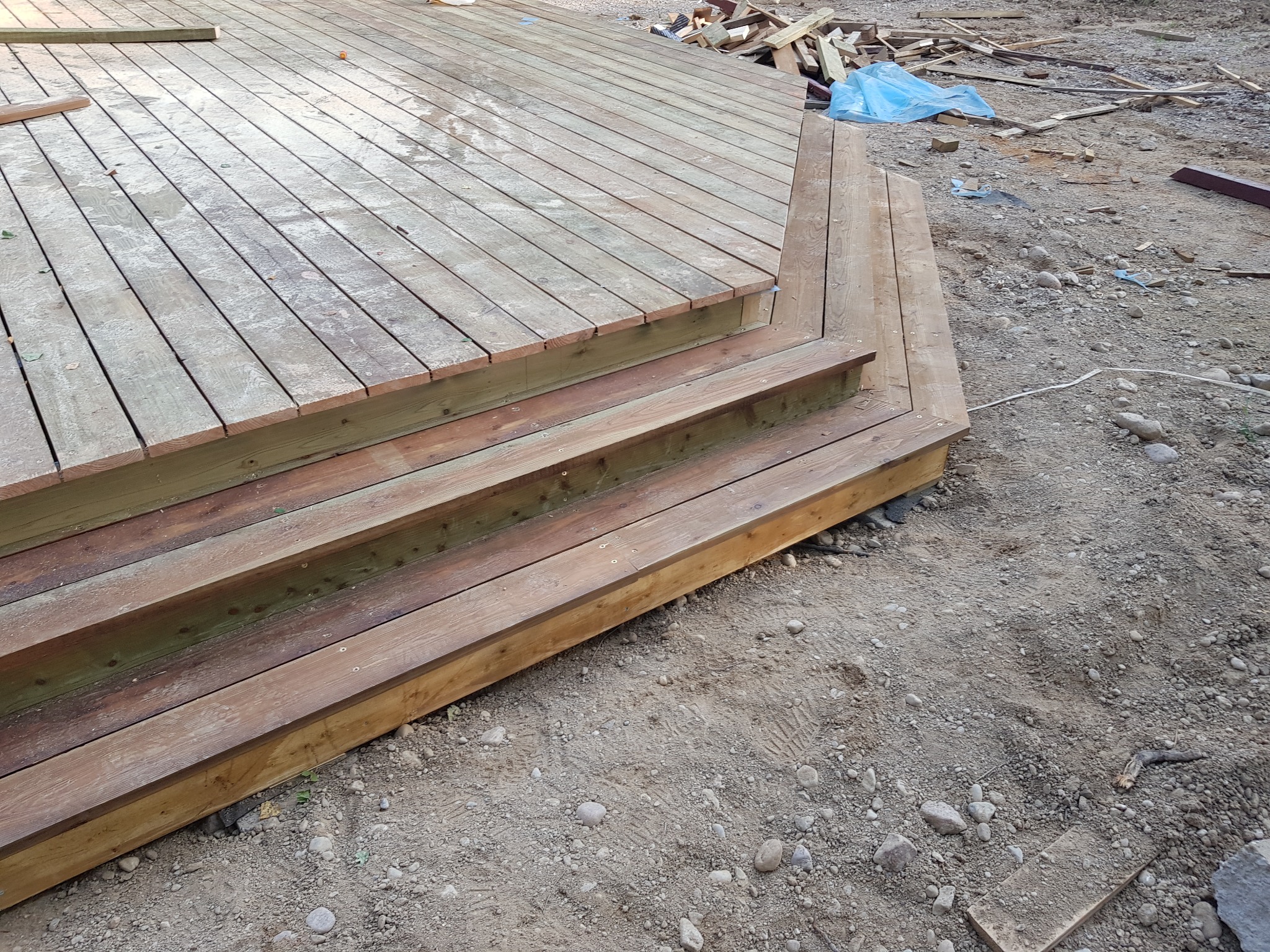 Drewniany taras z trzema schodkami w trakcie budowy, widoczne ślady pracy i resztki materiałów na ziemi.