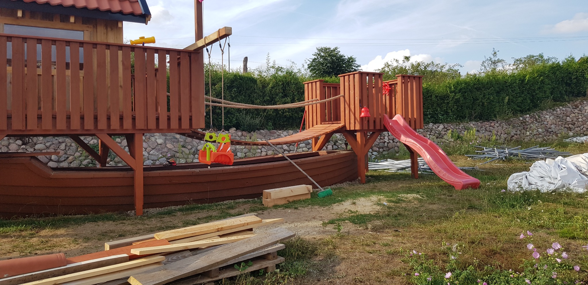 Drewniany plac zabaw w kształcie statku z huśtawką, zjeżdżalnią i mostkiem linowym, otoczony zielenią i kamiennym murem