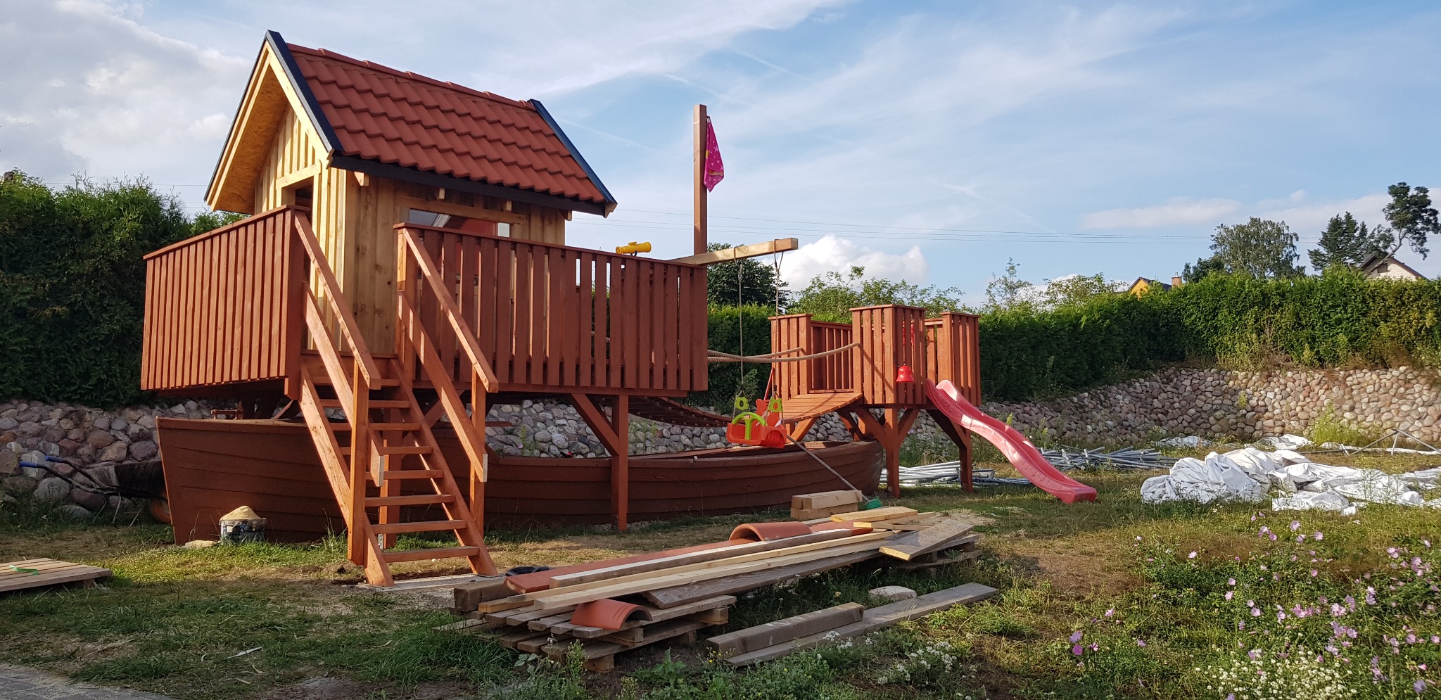 Drewniany plac zabaw w kształcie statku z domkiem, schodami, zjeżdżalnią i huśtawką, częściowo umieszczony na kamiennym podłożu, z elementami budowlanymi leżącymi obok, w tle zielony żywopłot.