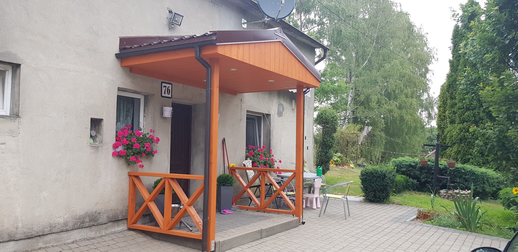 Zadaszone wejście do domu z drewnianą balustradą w kolorze pomarańczowym, ozdobione kwiatami w oknach, widok na ogród z tyłu.