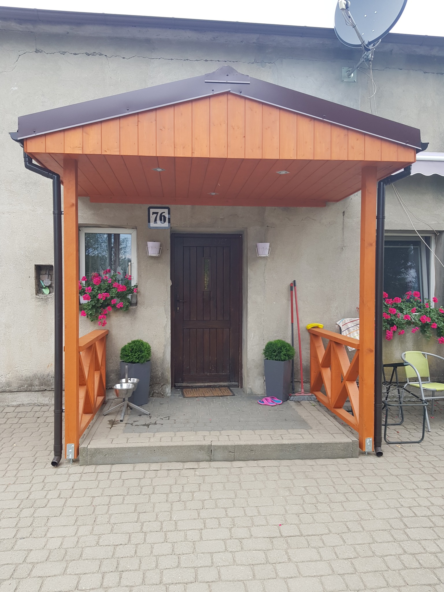 Zadaszone wejście do domu z drewnianą konstrukcją, numerem 76 nad drzwiami, kwiatami w oknach i roślinami w donicach po bokach.