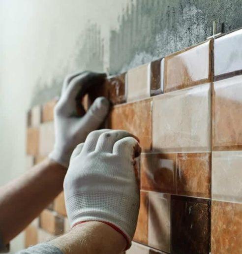 Ręce w białych rękawicach układają brązowo-beżowe płytki na ścianie w łazience, widoczne krzyżyki dystansowe i świeży klej.