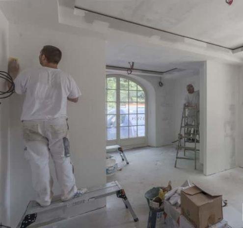 Pracownik w Częstochowie szlifuje ścianę gipsową po montażu płyt karton-gips, widoczny drugi pracownik na drabinie w tle i przygotowane narzędzia pracy.