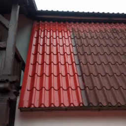 Częściowo wyczyszczony, czerwony dach z blachy falistej, pokazujący różnicę między umytą a brudną powierzchnią, widoczny fragment budynku z drewnianymi elementami.