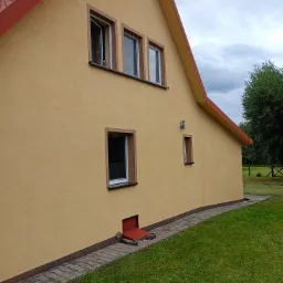 Żółta elewacja domu jednorodzinnego z czerwonym dachem i obramowaniami okien, widok z boku na trawnik i chodnik z kostki brukowej, jedno okno otwarte.