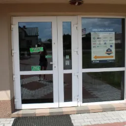 Wejście do biura z plakatem informującym o ofercie różnych towarzystw ubezpieczeniowych, widoczne szyldy firm ubezpieczeniowych.