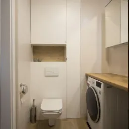 Jasna łazienka z toaletą wiszącą, pralką pod drewnianym blatem i szafką w zabudowie z półką na dekoracje.