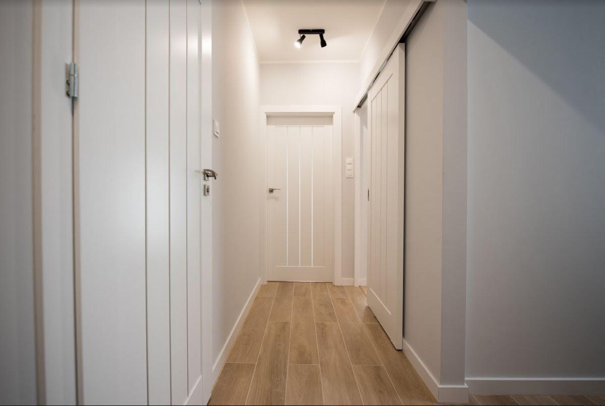 Jasny korytarz z drewnopodobną podłogą, białymi drzwiami i przesuwnymi drzwiami po prawej stronie, oświetlony dwoma reflektorami sufitowymi.