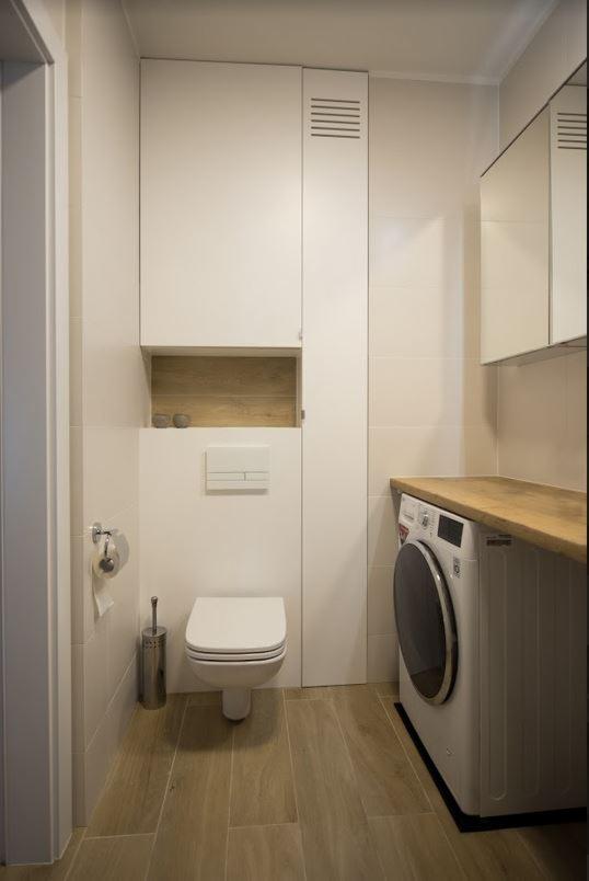 Jasna łazienka z toaletą wiszącą, pralką pod drewnianym blatem i szafką w zabudowie z półką na dekoracje.