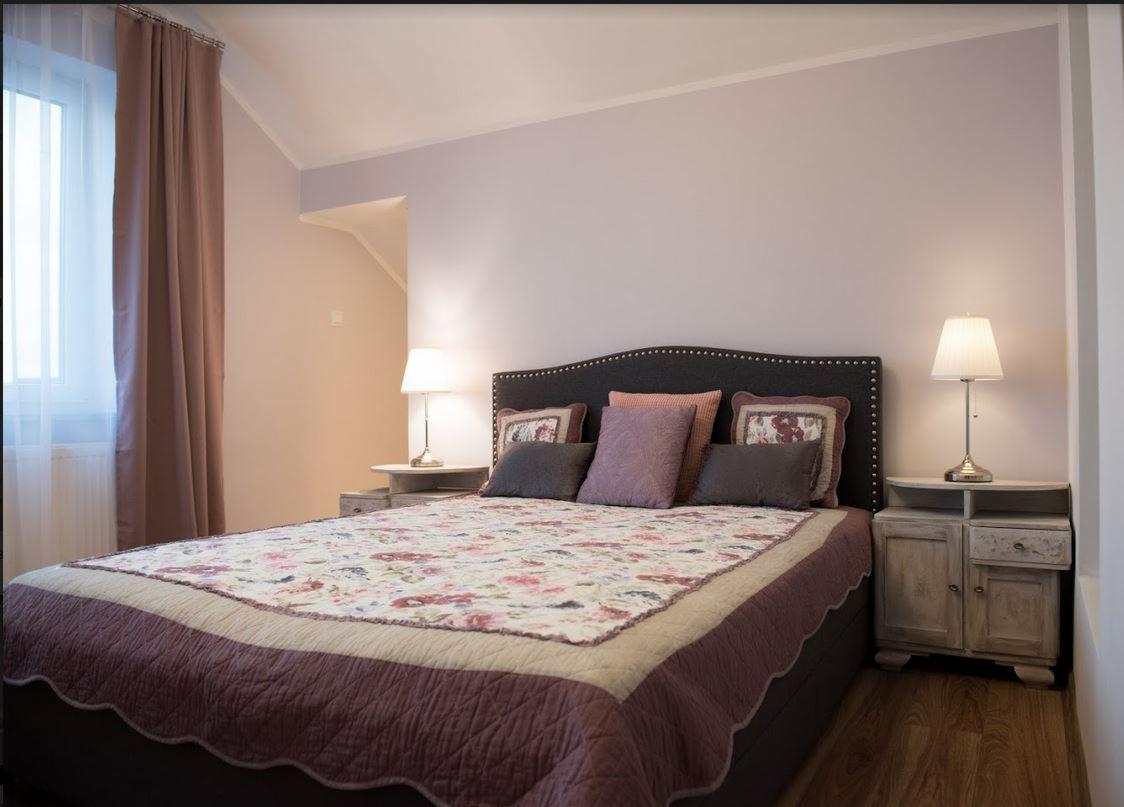 Sypialnia z łóżkiem z pikowanym zagłówkiem, kwiatową narzutą, szarobrązowymi poduszkami i lampkami nocnymi na drewnianych szafkach w stylu vintage.