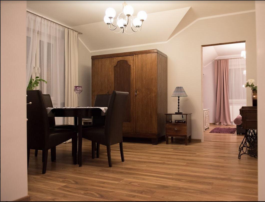 Jadalnia w ciepłych barwach z drewnianą podłogą, dużym, stylizowanym kredensem, ciemnymi krzesłami przy czarnym stole, oświetlona lampą sufitową i lampką nocną na stoliku.